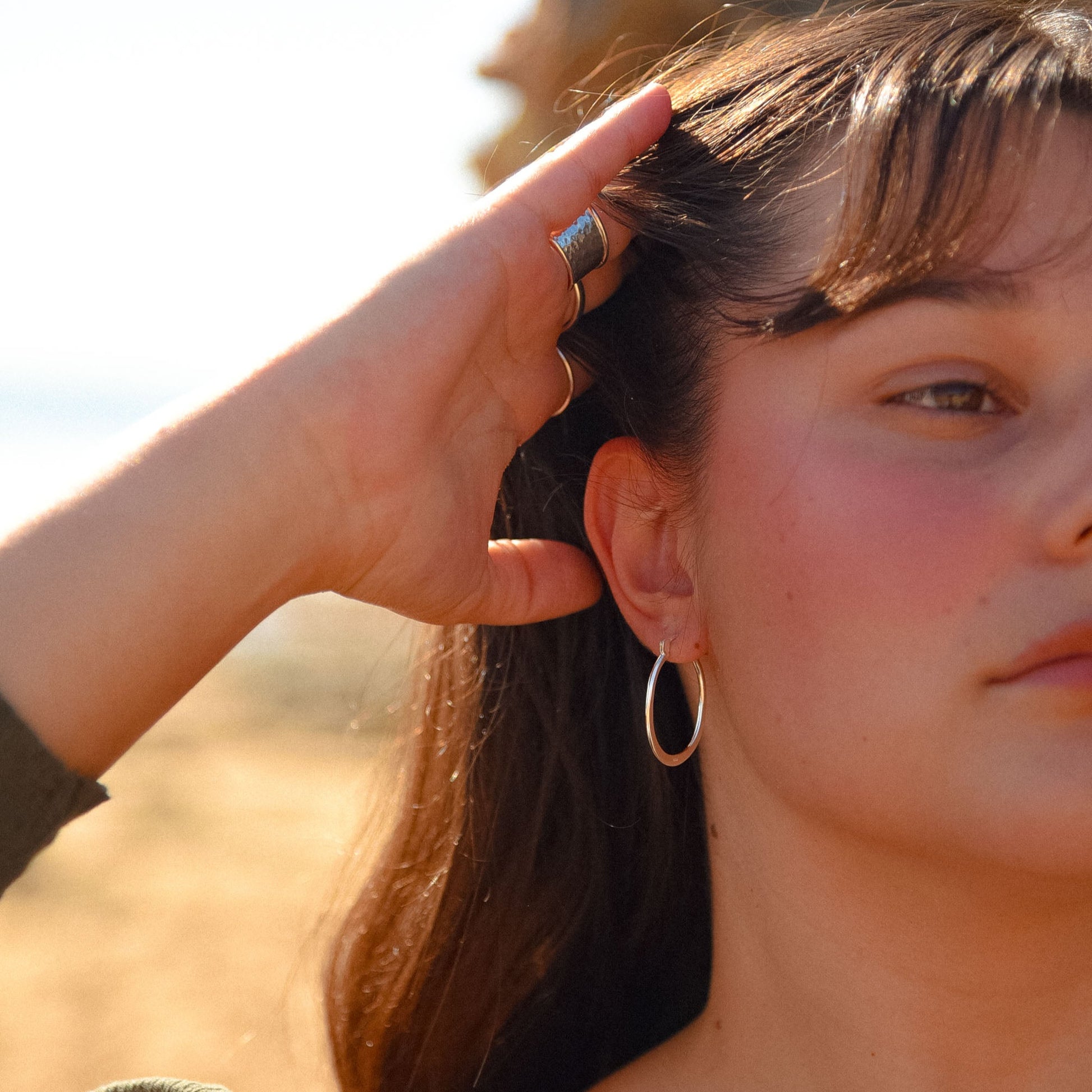 Woman adjusting her hair with a blurred natural background. wearing the ocean haze hoops which are a classic thin look hoop with a unique oval shape. 
These hoops are salt water safe
