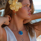 Woman with a yellow flower in her hair and a blue pendant necklace, standing on a beach.
